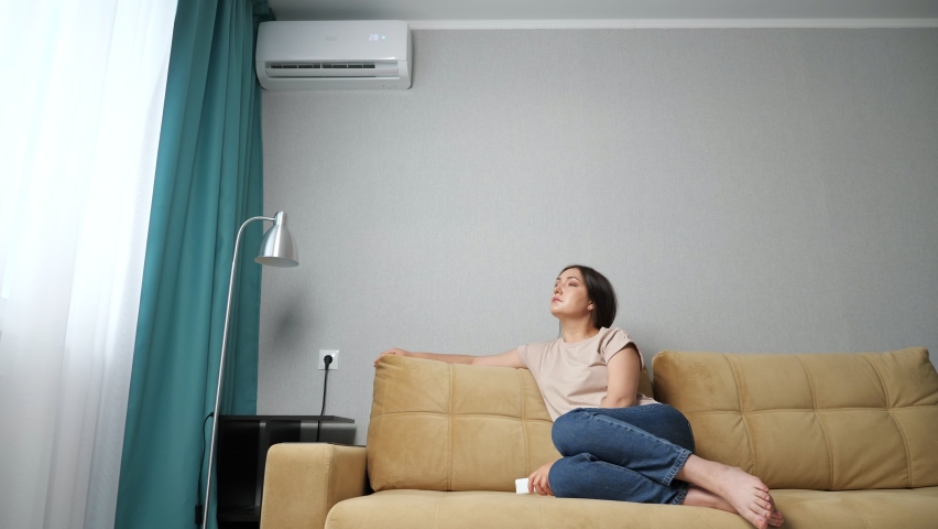 brunette woman turns on the air conditioner while sitting on the couch. Royalty-Free Stock Footage #1074949172