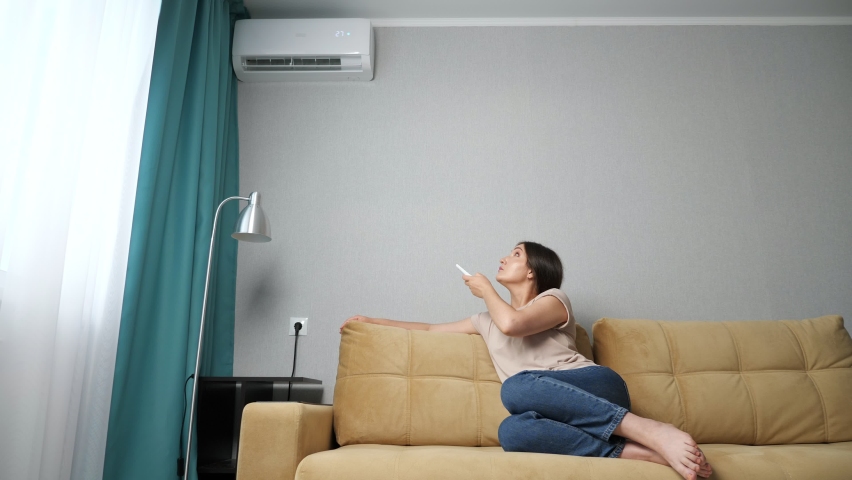 Brunette woman turns on the air conditioner while sitting on the couch. | Shutterstock HD Video #1074949172