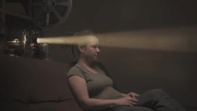 Woman watching scary film on old movie projector sitting on couch in dark room.