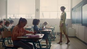 Multi ethnic children doing online test with digital tablets in school classroom. Serious schoolteacher walking between desks with students. Female teacher giving information to pupils in school class - Powered by Shutterstock - Get 15% off with code: PIKWIZARD15