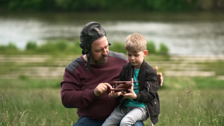 A bearded father and son are playing with a wooden toy airplane in nature. A man puts a pilot