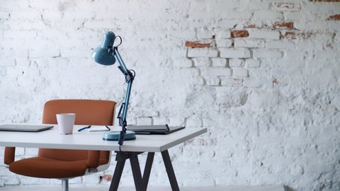 Bright contemporary modern office work space with white brick wall background orange chair blue lamp coffee cup laptop computer monitor keyboard pencils and notes