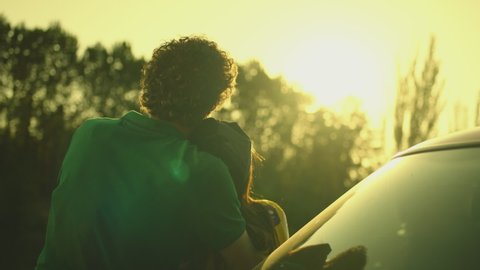 Back rear view of young couple sitting in auto car hood looking at sunset and beautiful nature , hugging , caressing each other . Boy and girl standing by the machine , holding hands  . Slow motion