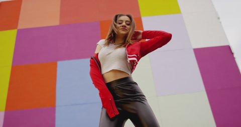 Beautiful , stylish , young girl with blond hair and beautiful body in Leather tights dancing , moving , smiling and looking at camera with happy emotions . Slow motion . Colorful wall background