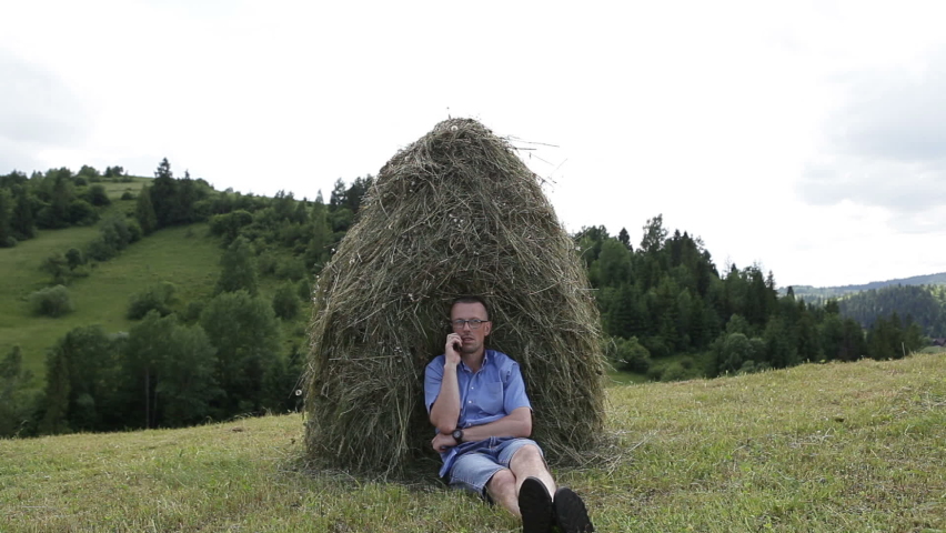 A man is talking on a cell phone near a haystack. A 45-year-old man is resting in a village high in the mountains from the bustle of the city.
