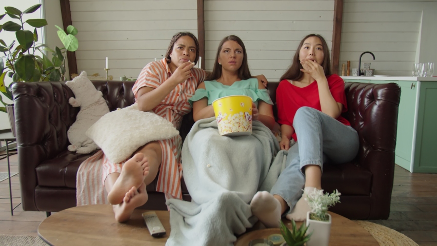 Happy diverse group of women watching sports event on TV online using app sitting on sofa at home, eating popcorn, throwing arms up, giving high five in excitement celebrating win of team.