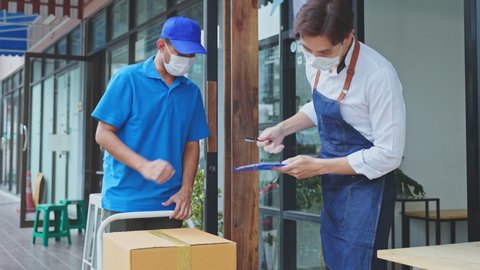 Asian Postman deliver food ingredient box to restaurant waiter in cafe. Attractive barista worker wear mask due to Covid-19, sign for parcel and carry box from deliverly in blue uniform to coffeehouse