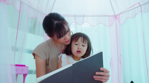Asian mom read storytale to young baby girl daughter in tent at home. Beautiful mother play education game picture book with happy little cute kid to develop skill. Parenting activity in house concept
