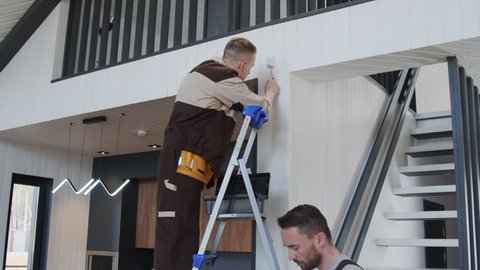 Tilt up slowmo shot of handyman writing on clipboard while electrician in uniform standing on ladder and inspecting smart home device or sensor