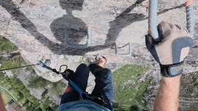 Male hiker ascends vertical via ferrata route. He clips cables and follows metal rungs while doing so
