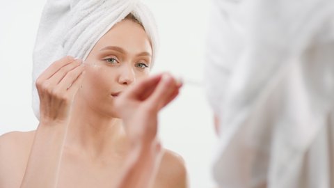 Good-looking fit European young woman wrapped in a towel applies skin care serum on her face using a dropper and smiles wide in front of the mirror on white background | Skin care serum commercial