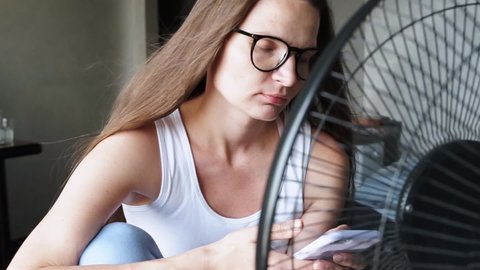 Tired overheated young hipster woman wear glasses suffering from high temperature in home office sitting in front cooling fan, ventilator. Hot weather. People texting phone check social media.