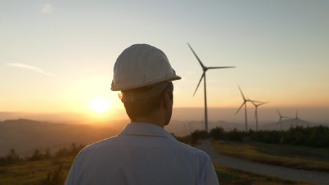 Cinematic shot of young engineer putting security helmet on satisfied with his job on background of wind mills with at sunset.Concept: renewable energy, technology, electricity, service, green, future