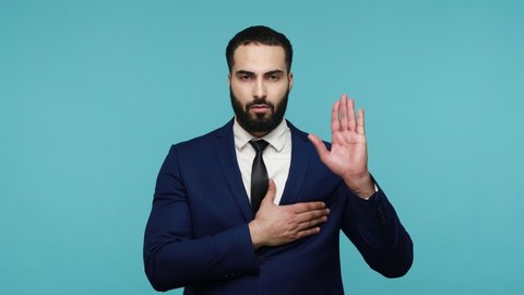 I swear! Responsible serious young businessman in black suit giving promise with raised hand and nodding approvingly, honest guy making promise oath. Indoor studio shot isolated on blue background.