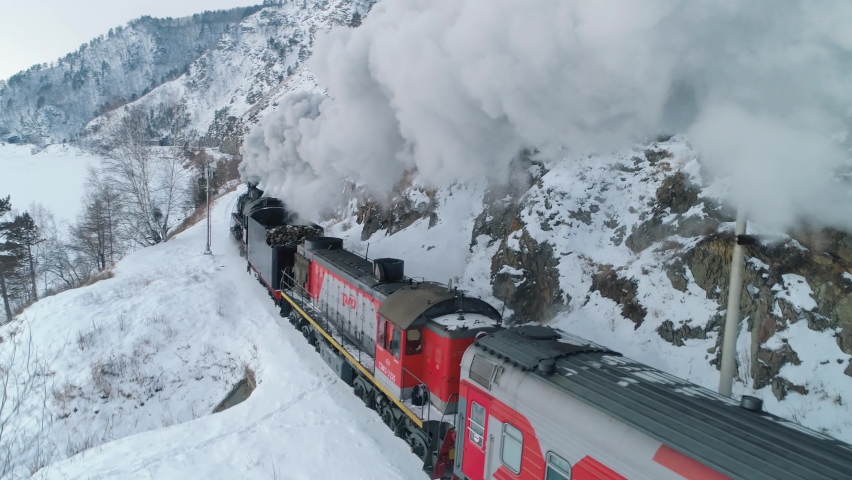 Russia Siberia 20 January 2020: Aerial old steam locomotive, engine rides on railroad releases thick clouds of steam from pipe. Winter snowy nature. Historical Soviet Union Trans-Siberian railway film