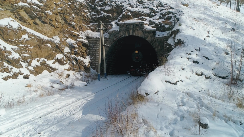 Aerial close to old steam locomotive engine leaving dark tonel in slow motion, historic train traveling on railroad railway. Steam puffs of smoke from chimney. Trans-Siberian Russia Circum-Baikal film