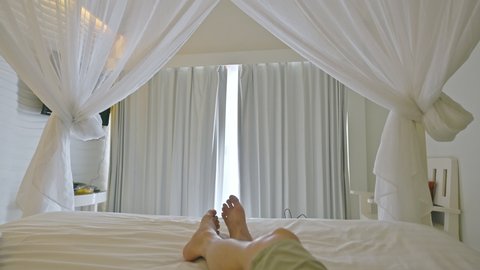 A man resting and wiggling feet comfortably in the bed with mosquito net and white curtain closed in the villa at vacation