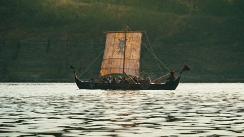 Vikings Sail on an Old Ship with a Lowered Sail on a Calmt River Against the Backdrop of Rocky Mountains. Concept on the theme of the Vikings and the early Middle Ages.
