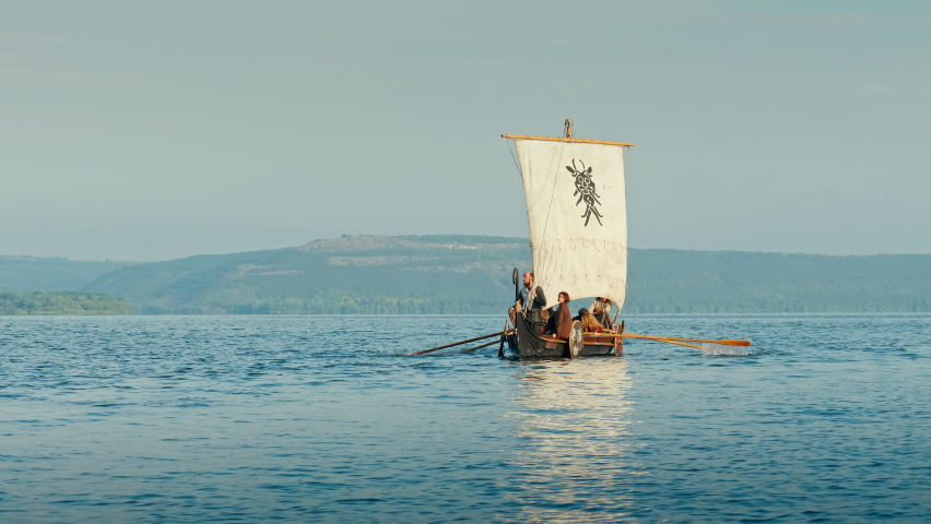 Vikings Sail on an Old Ship with a Raised Sail on a Calm River Against the Backdrop of a Rocky Coast. The Men Row the Oars Diligently Towards Adventure. Medieval Reconstruction. Royalty-Free Stock Footage #1074984068