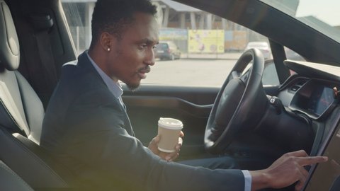 Side view of focused african man sitting on driver's seat with cup of coffee and using dashboard for navigation. Businessman in formal wear taking stop for checking direction. Man sitting in car
