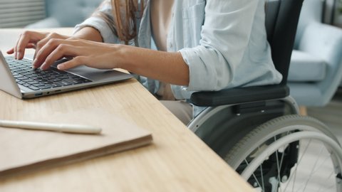 Slow motion of female student learning online using laptop typing sitting in wheelchair in apartment busy with distant education. Technology and disability concept.