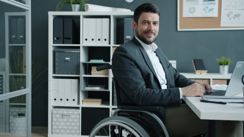 Portrait of disabled employee sitting in wheelchair looking at camera and smiling in workplace. Ambitious people with special needs and job concept. Royalty-Free Stock Footage #1074987407
