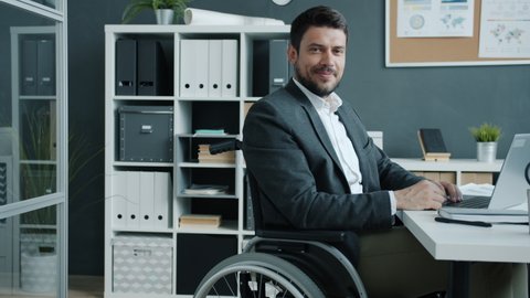 Portrait of disabled employee sitting in wheelchair looking at camera and smiling in workplace. Ambitious people with special needs and job concept.