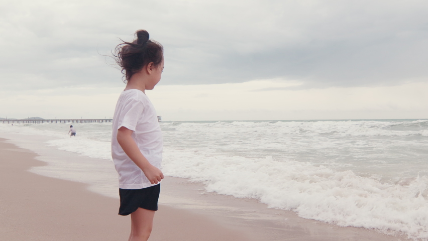 4K of adorable 5 years old asian girl with black hair is running with happy smile and enjoyment near ocean coast seeing strong wave and horizon for sunset which is relaxing holiday for kid and family.