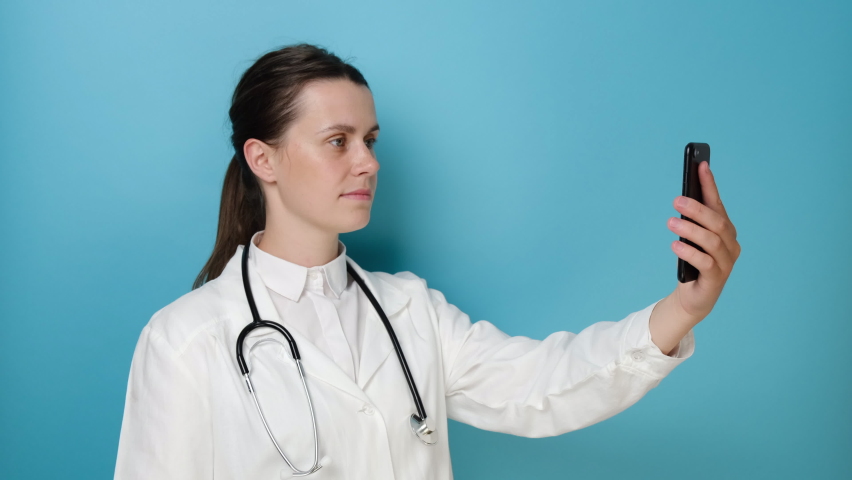 Smiling young female doctor holding phone talk to patient make telemedicine online conference video call, posing over blue wall. Gp remote consultation telemedicine virtual mobile health care chat app Royalty-Free Stock Footage #1074993482