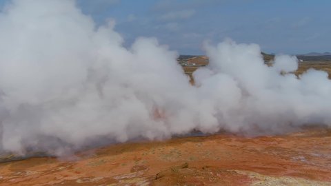 ICELAND - CIRCA 2020s - Aerial past steaming hot fumeroles in a geothermal aera reveals a geothermal power plant in the distance.