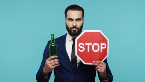 Do not drive drunk. Bearded businessman showing empty alcoholic beverage bottle and stop sign, warning and worrying, prefers healthy lifestyle. Indoor studio shot isolated on blue background.