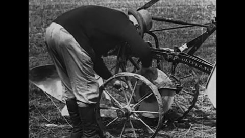 CIRCA 1920 - A farmer puts wires on his tractor to work as a trash guard while he plows a corn field.