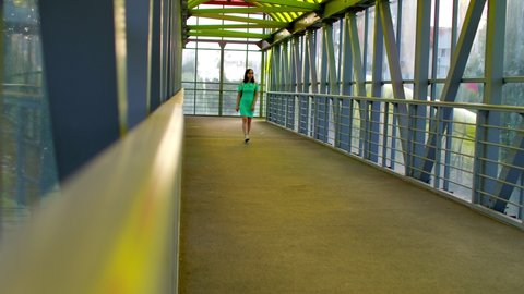 Front view of young woman in dress going pedestrian crossing. Brunette walking along overground pedestrian in summertime. Concept of safe transition above highway.
