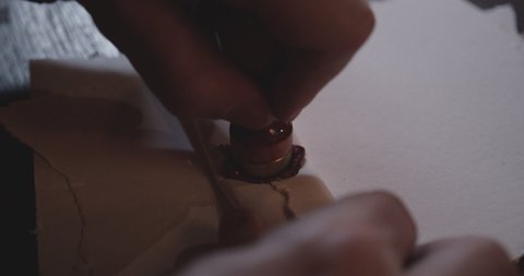 An angled close-up shot of a person's hands sealing an old parchment paper putting pressure on the bronze-colored stamp as it seals the paper with its red wax showing the letter insignia