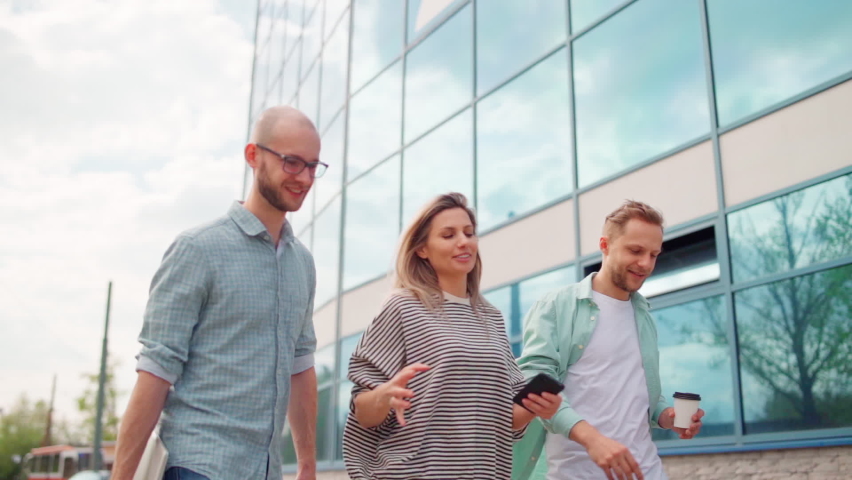Tracking slow motion shot of group of three business people, men with laptop and coffee and woman with cell phone, walking outside near office building, talking cheerfully and laughing - Powered by Shutterstock - Get 15% off with code: PIKWIZARD15