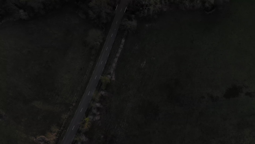 Drone flies over a small road at dusk as cars drive down a two-lane highway. cars, field and trees in a Aerial dark moody shot of road. car driving down a dark rural road outside the city at night.