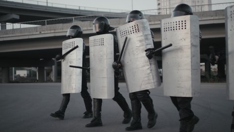 Slowmo shot of group of riot police officers in full gear walking and beating shields with their batons while trying to cause fear in protesters