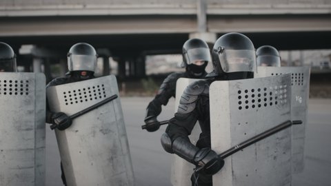 Slowmo shot of unrecognizable riot police officers in full gear beating their shields with batons while trying to intimidate protesters