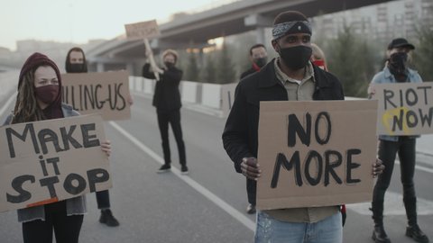 Handheld slowmo shot of young people in face masks blocking road and protesting with signs