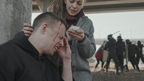 Close up slowmo shot of young woman with dreadlocks helping wounded man while riot police pushing back protesters in background