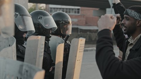 Slowmo tracking shot of riot police officers in full gear holding shields and standing in row while young people chanting and protesting