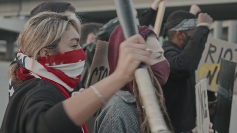 Slowmo tracking shot of masked young people with signs chanting and protesting before group of intimidating riot police officers with shields