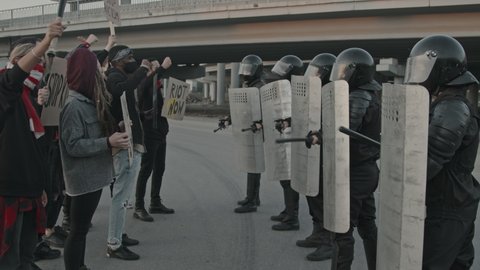Tracking slowmo shot of young people in face masks holding signs and protesting in front of riot police officers beating shields with batons and blocking way