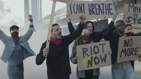 Handheld slowmo tracking shot of diverse group of young people with smoke bomb and signs walking and chanting while protesting outside