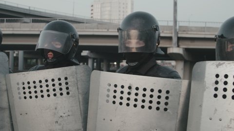 Slowmo tracking shot of riot police officers in masks and helmets holding shields and blocking street during protest