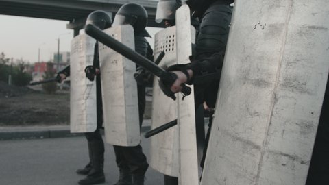 Tilt up slowmo shot of unrecognizable riot police officers in full gear blocking street and beating shields with batons