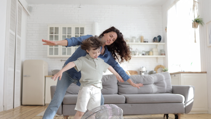 Family bonding at home: playful mother and preschool kid have fun with big indoor fan blow cool wind. Young single parent and child weekend indoor engagement. Cheerful mum enjoy leisure time with son | Shutterstock HD Video #1075012823