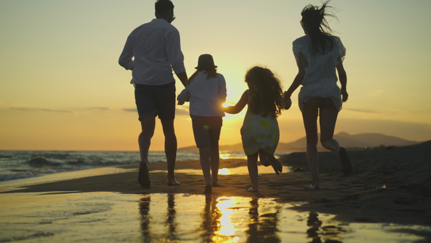 Tracking back view footage of young family holding hands and running on beach against setting sun. Summer evening at seaside. Royalty-Free Stock Footage #1075013246