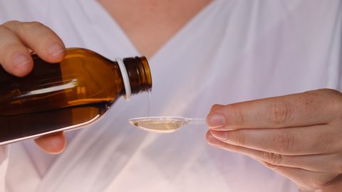 Cough syrup is poured into transparent spoon. woman doctor in white coat from brown transparent bottle pours medical drug liquid into plastic spoon. Treatment of a disease, sick person.