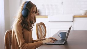 Teenage caucasian girl with bluetooth and wireless headphones using laptop while chatting over smartphone with coffee at home